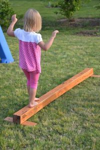 A young child with blonde hair balances on a wooden beam in a grassy area, with a blue object partially visible to the left.