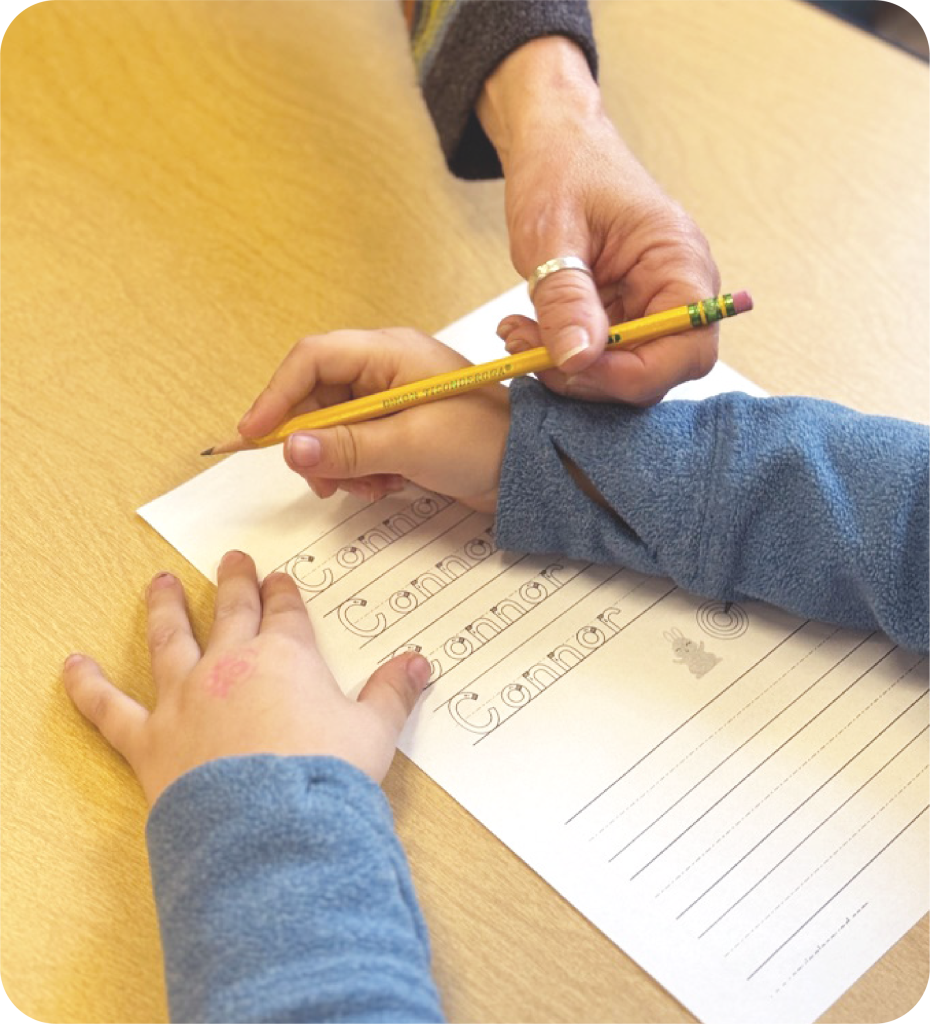 A child practices writing "Connor" on paper with an adult guiding their hand with a pencil.