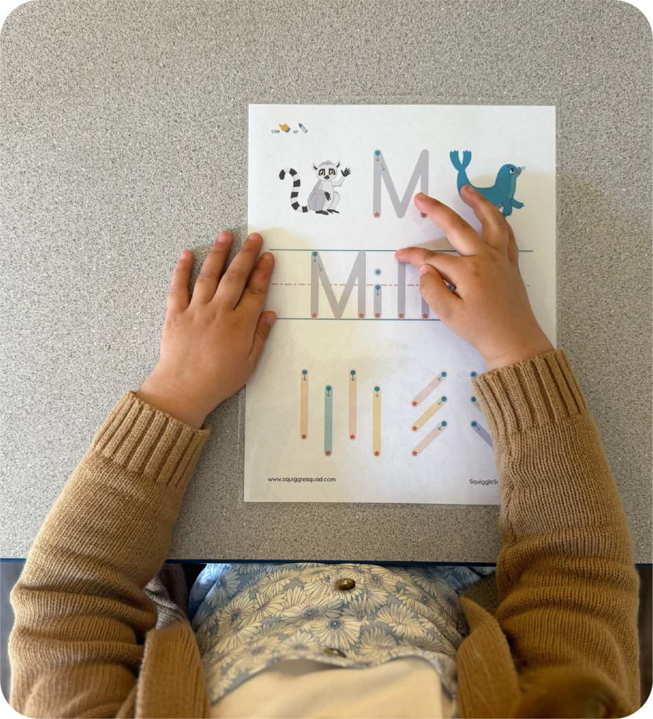 Child tracing letters on a worksheet with animal illustrations, wearing a brown sweater and floral shirt.