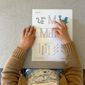 A child traces the letter "M" on an animal-themed worksheet using their fingers to follow the dotted lines, part of the Name Generator - Yearly School Access Pass activities.