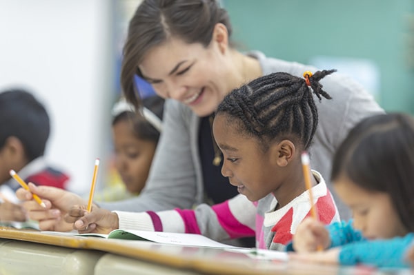 A teacher smiles as she helps a student with dark hair in braids. Other children are focused on their work at their desks.