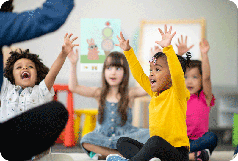 Four young children sit cross-legged indoors with their arms raised, appearing engaged in an activity led by an adult. Classroom materials and colorful furniture are visible in the background.