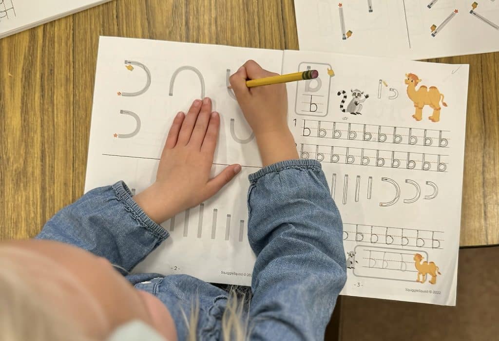 A child writes with a pencil on a worksheet featuring letters, lines, and animal illustrations on a wooden table.