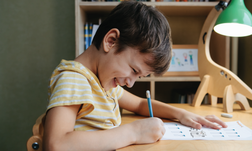 A young child sits at a desk, smiling while writing on a worksheet with a pencil. A lamp and shelves with books are in the background.
