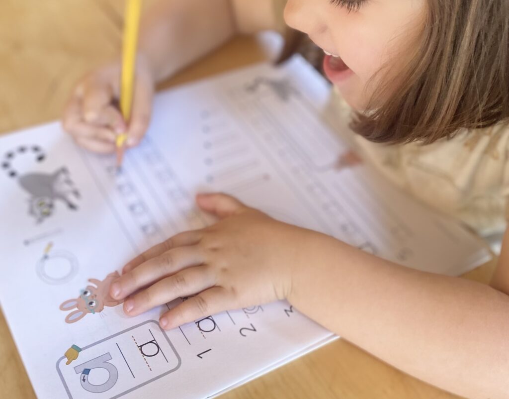 A child writes with a pencil on a worksheet featuring numbers and animal illustrations at a wooden table.